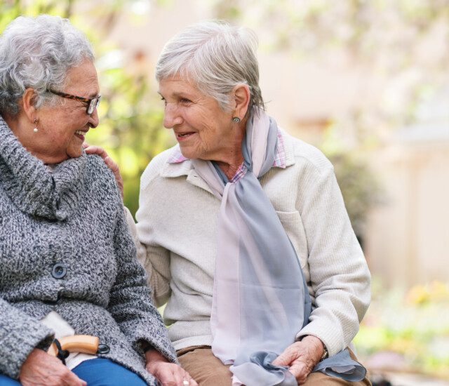 Two elderly women sitting on bench in park smiling happy life long friends enjoying retirement