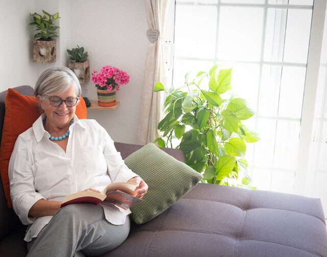 Senior pretty lady resting on the couch reading a book and drinking a fruit juice. Corner with plants and decorations against a white wall. Bright light from the window