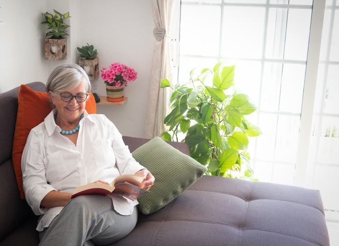 Senior pretty lady resting on the couch reading a book and drinking a fruit juice. Corner with plants and decorations against a white wall. Bright light from the window